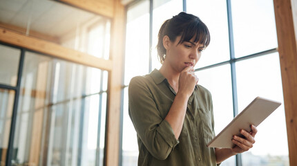 Young professional woman standing in a modern office, looking worried while analyzing data or reading something on her tablet.