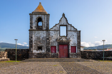 Ruins of the Old Church of S&atilde;o Mateus on Terceira Island, Azores, Portugal, showcasing historic stone architecture, red wooden doors and a cobblestone square under clear blue sky