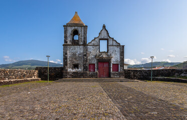 Ruins of the Old Church of S&atilde;o Mateus on Terceira Island, Azores, Portugal, showcasing historic stone architecture, red wooden doors and a cobblestone square under clear blue sky