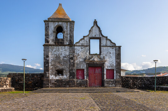 Ruins of the Old Church of S&atilde;o Mateus on Terceira Island, Azores, Portugal, showcasing historic stone architecture, red wooden doors and a cobblestone square under clear blue sky