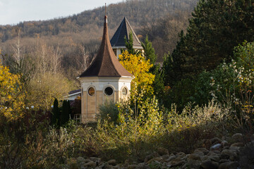 Country house is located on banks of mountain river. Octagonal tower with pointed roof and round windows, partially hidden by lush foliage, rises above autumn trees and is surrounded by cliffs