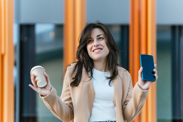 Cheerful businesswoman holding smartphone and coffee on trip