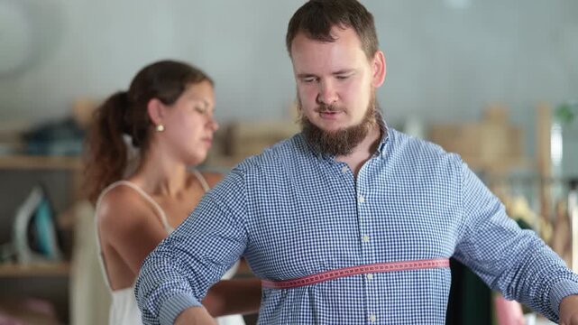 Young seamstress skillfully taking measurements for male client in modest sewing studio, preparing for custom-made clothing order