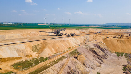 High-angle drone shot of a massive lignite mine landscape. Industrial scene featuring heavy mining machinery, green fields, wind turbines and thermal power station under a clear sky