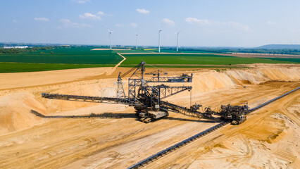 High-angle drone shot of a massive lignite mine landscape. Industrial scene featuring heavy mining machinery, green fields, wind turbines and thermal power station under a clear sky