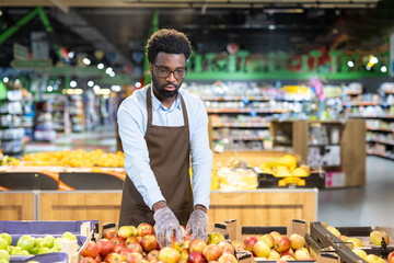 Young african american man wearing an apron and gloves arranging fresh red and green apples on a display stand in the produce section of a grocery store, ensuring quality and presentation