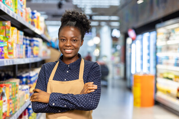 African american woman in a blue shirt and apron, arms crossed, smiling at the camera while standing confidentially in a grocery store aisle with shelves filled with products
