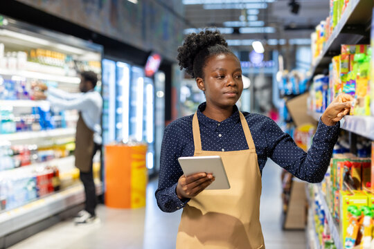 Young african american woman in an apron and checkered shirt uses a digital tablet to check and organize products on grocery store shelves, focused on inventory management and efficient restocking