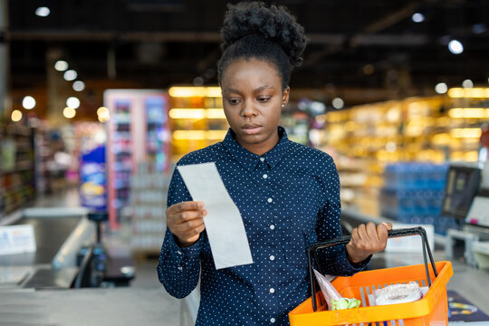 African american woman in supermarket aisle holds shopping basket and reads a long receipt with a worried expression, signaling rising grocery costs and budget strain