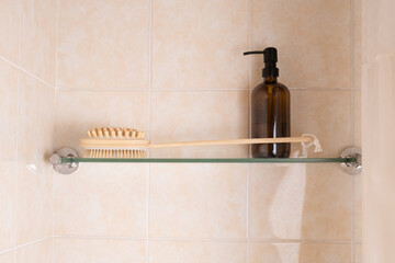 Wooden bath brush and refillable dispenser bottle arranged on glass shelf