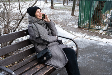 woman bundled in a warm coat sitting on park bench and talking on smartphone