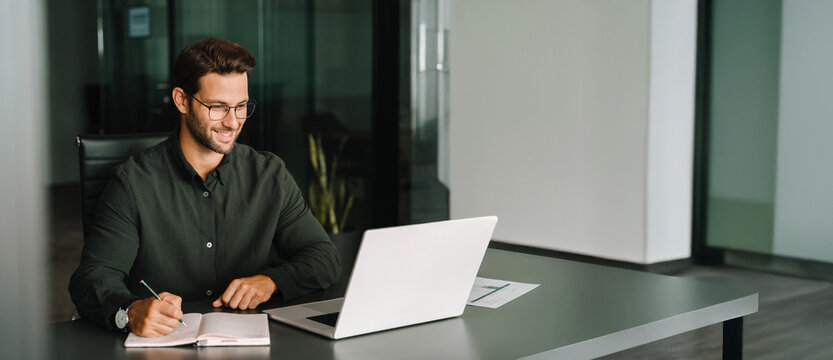 Middle-age European man using laptop computer for business studying, watching virtual webinar training writing notes. Focused young Latin businessman student working in office. Web banner, copy space