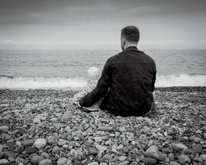 B&W back view of a father and child sitting on a pebble beach, looking at the sea. Concept of family, peace, and bonding.