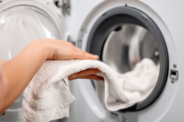 Hand of a person placing a soft white towel into a modern washing machine, showcasing the laundry process in a bright, clean laundry room environment with focus on cleanliness