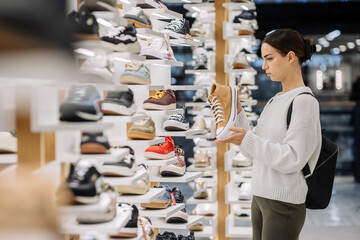 Young woman examining stylish sneaker in modern shoe store, surrounded by various footwear displays, showcasing contemporary fashion trends and shopping experience