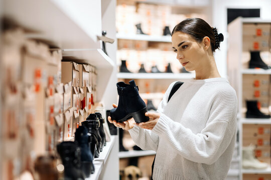 Young woman with dark hair, wearing a cozy sweater, examines stylish black ankle boots in a modern shoe store, surrounded by neatly arranged footwear and boxes