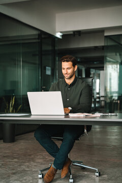 Young professional specialist latin hispanic business man working on laptop pc sit at desk in office. European businessman entrepreneur using computer technology for work online. Full body, vertical