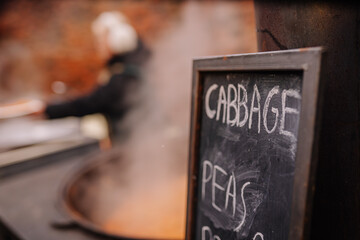 Freshly prepared cabbage and peas displayed on a chalkboard sign, with steam rising from a large cooking pot in the background, showcasing a vibrant culinary scene