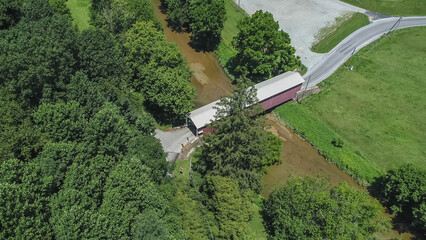 A view of a covered bridge located in a rural setting with trees surrounding it. The water nearby reflects the greenery. A gravel road runs next to the area, showing the landscape's features.