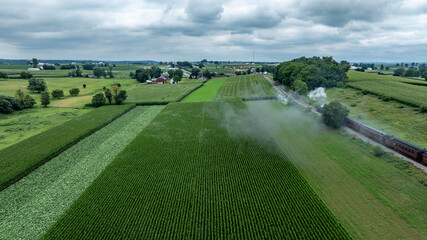 A steam train moves along the tracks, surrounded by vibrant green fields and farmland. The scenery features rolling hills, farms, and cloudy skies, creating a peaceful rural setting.
