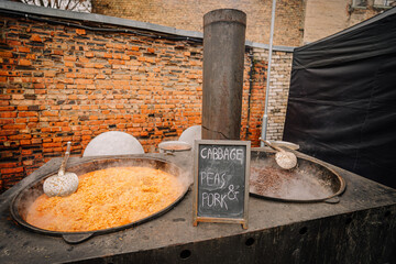 Large metal cooking pots filled with vibrant cabbage, peas, and pork, steaming in an outdoor setting, showcasing traditional culinary preparation and communal dining experience