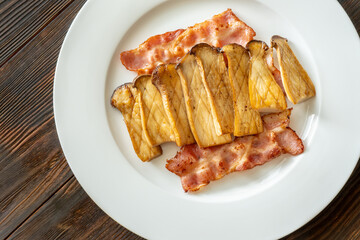 Fried eryngii mushrooms served with crispy bacon slices on white plate