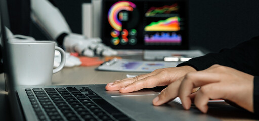 A modern workplace scene shows a person using a laptop with a coffee cup nearby, while a robotic arm interacts with vibrant data visualizations in the background.Noogenesis