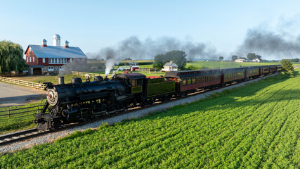 A steam train runs along a track beside green fields and a barn. The sky is clear and the sun is...