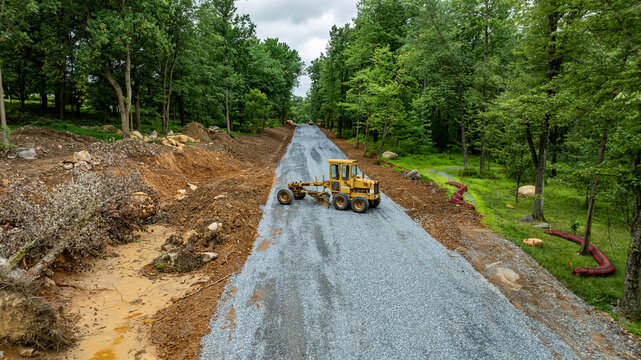 Heavy machinery is working on a rural road construction site. Gravel is laid down on the road. Trees and dirt piles surround the area. Workers are present in the background.
