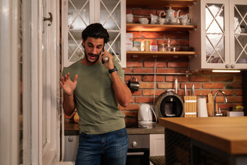 Young man in a modern kitchen on an intense phone call, gesturing with his hand and showing stress and frustration while discussing a serious problem on his smartphone