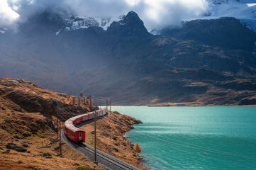 Aerial view of red Bernina Express train, railroad, turquoise lake in the Swiss Alps, rocky hills and dramatic cloudy sky at sunset in autumn. Alpine mountains in snow. Top view of train in fall