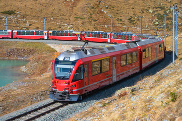 Red modern train is moving along railway in swiss alps near mountain lake on sunny day in autumn in Switzarland. Bernina Express train, orange grass, hills at sunset in fall. Railway travel. Railroad