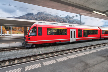 Red modern passenger train on mountain railway station in Swiss Alps in autumn. High-speed train, railway platform and cloudy sky. Bernina Express. Rail transport. Station in St. Moritz, Switzerland