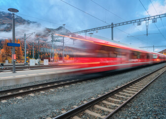 Naklejka premium Blurred red passenger train passing mountain railway station in Swiss Alps at dusk. Moving high-speed train. St. Moritz, Switzerland. Bernina Express. Railway platform and cloudy sky. Rail transport