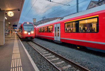 Red modern trains on mountain railway station in Swiss Alps in autumn at night. Speed trains with lights, railroad, railway platform in fall at dusk in St. Moritz, Switzerland. Bernina Express