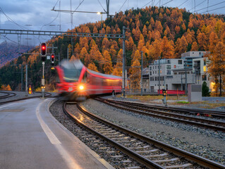 Blurred red passenger train passing mountain railway station during rain in autumn Swiss Alps at night. Moving speed train at dusk. St Moritz, Switzerland. Bernina Express. Railway platform and lights