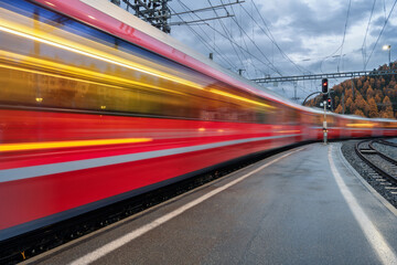 Blurred red passenger train passing mountain railway station during rain in Swiss Alps at night. Moving speed train at dusk. St. Moritz, Switzerland. Bernina Express.  Railway platform and lights.