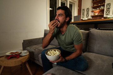 Young man relaxing on sofa at home in the evening, smiling while watching tv and eating popcorn...