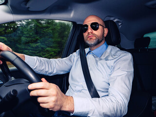 Male driver in his 50s sitting in a car in a green forest area. Bold male in light blue shirt with confident look. Mature driver insurance.