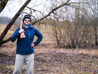 A man in a green jacket and hat is holding a rifle. He is smiling and looks confident handling the weapon. Hunter holding light small caliber gun. Grey winter snowless background.