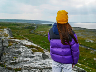 A girl is wearing a blue jacket and a yellow hat stands on a rocky hill overlooking the ocean. The scene is serene and peaceful, with the woman posing for a photo. Burren area, Ireland. Travel 