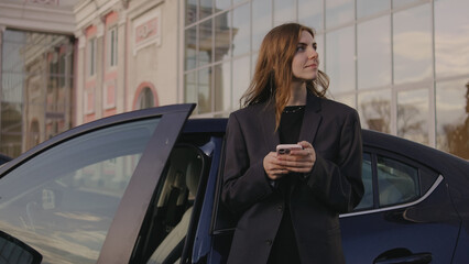 Woman stands next to a car, holding a phone in her hand while looking away from the vehicle at a building in the background in an outdoor setting © tol_u4f