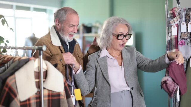 Mature husband and wife stand in a clothing boutique and choose underwear. Elderly couple buying a bra set in a lingerie store. High quality 4k footage
