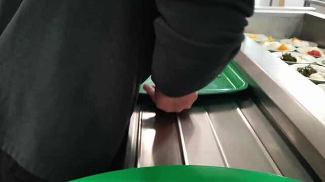 Customers slide trays along metal rails to select lunch dishes in a busy public dining room, showing a self-service cafeteria line and diverse meal options in high-quality 4k resolution.
