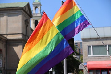Two large rainbow flags displayed outdoors against city architecture and blue sky