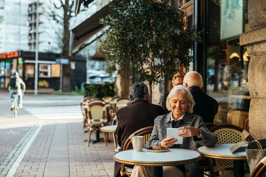 Smiling senior woman wearing wireless in-ear headphones using tablet PC while sitting in sidewalk cafe