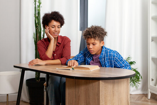 African American woman and her son read book and do homework together at home, mother helping teenager study