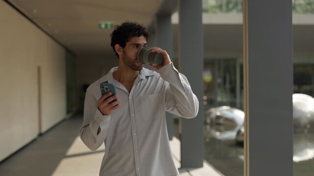 Portrait of Hispanic man with smartphone drinks coffee at business center slow motion. Handsome Latin male in loose shirt texts on cellphone in modern city. Morning routine