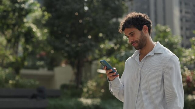 Portrait of happy Hispanic man texts on smartphone and drinks walks along sunny street slow motion. Bearded Latin guy in shirt sends message online via mobile phone in summer city