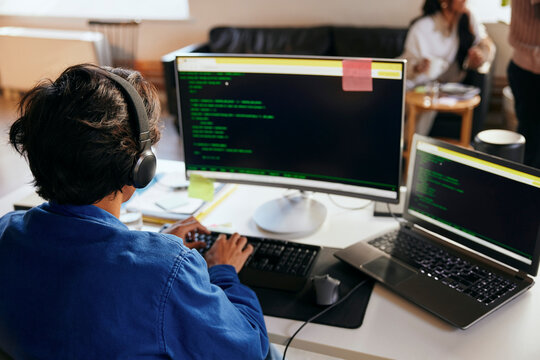 Rear view of young male programmer wearing wireless headphones coding on desktop PC in office